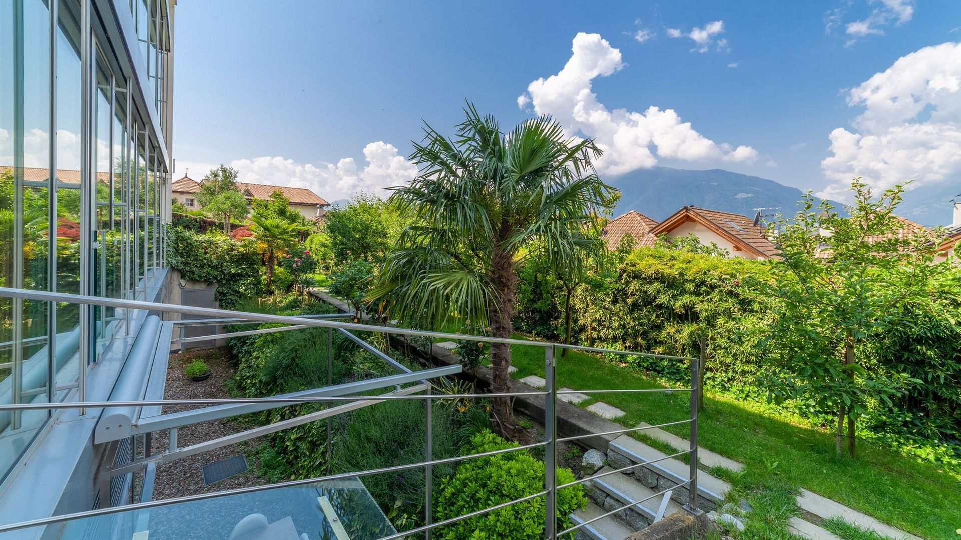 Balkon mit Glasfassade und Geländer, Blick auf grünen Garten mit Palme, Häusern und Bergen unter blauem Himmel. Balkon mit Glasfassade und Geländer, Blick auf grünen Garten mit Palme, Häusern und Bergen unter blauem Himmel.