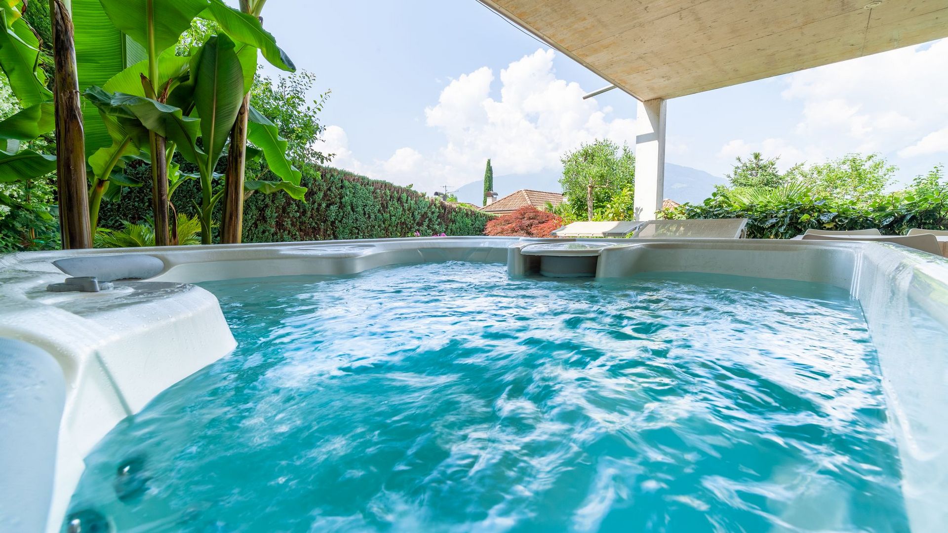 Sprudelnder Außenwhirlpool unter Überdachung, umgeben von grünen Pflanzen und Gartenblick. Sprudelnder Außenwhirlpool unter Überdachung, umgeben von grünen Pflanzen und Gartenblick.