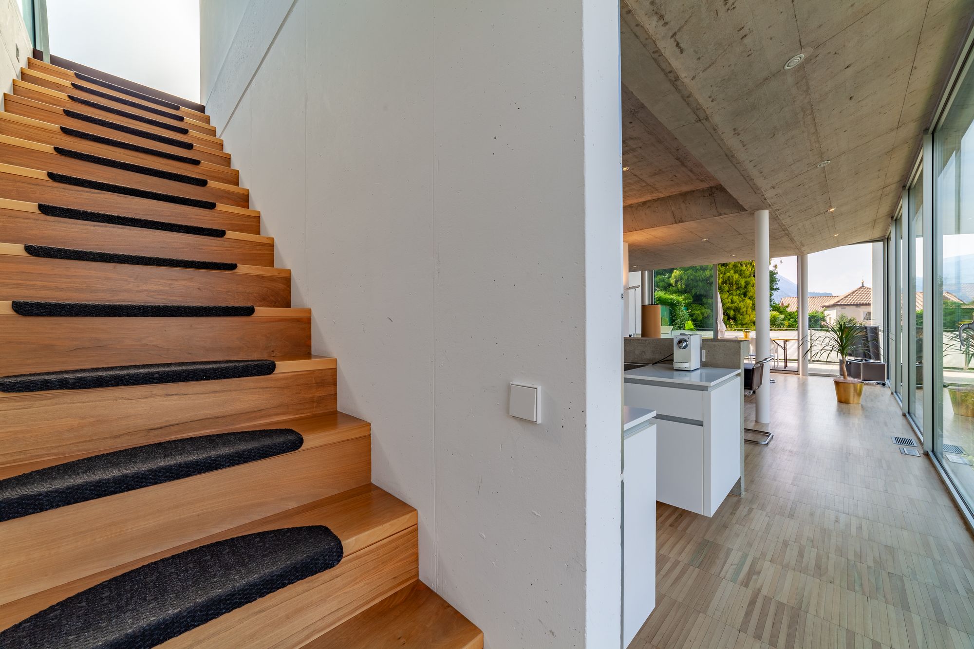 Holztreppe mit schwarzen Antirutschstreifen neben offenem, lichtdurchflutetem Wohnbereich mit großen Fenstern Holztreppe mit schwarzen Antirutschstreifen neben offenem, lichtdurchflutetem Wohnbereich mit großen Fenstern