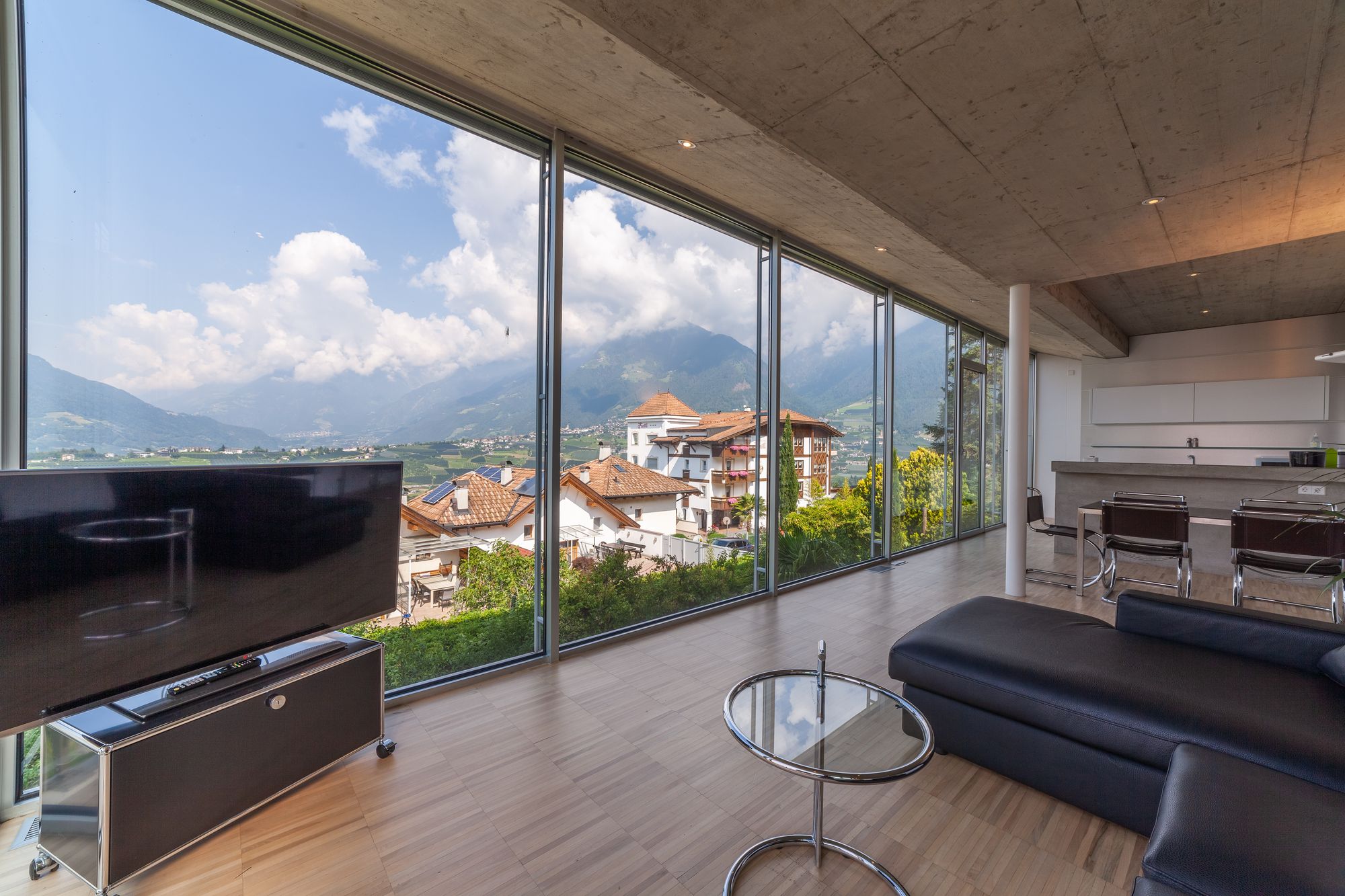 Modernes Wohnzimmer mit bodentiefen Fenstern, Blick auf Dächer und Berge, Couch und Fernseher Modernes Wohnzimmer mit bodentiefen Fenstern, Blick auf Dächer und Berge, Couch und Fernseher