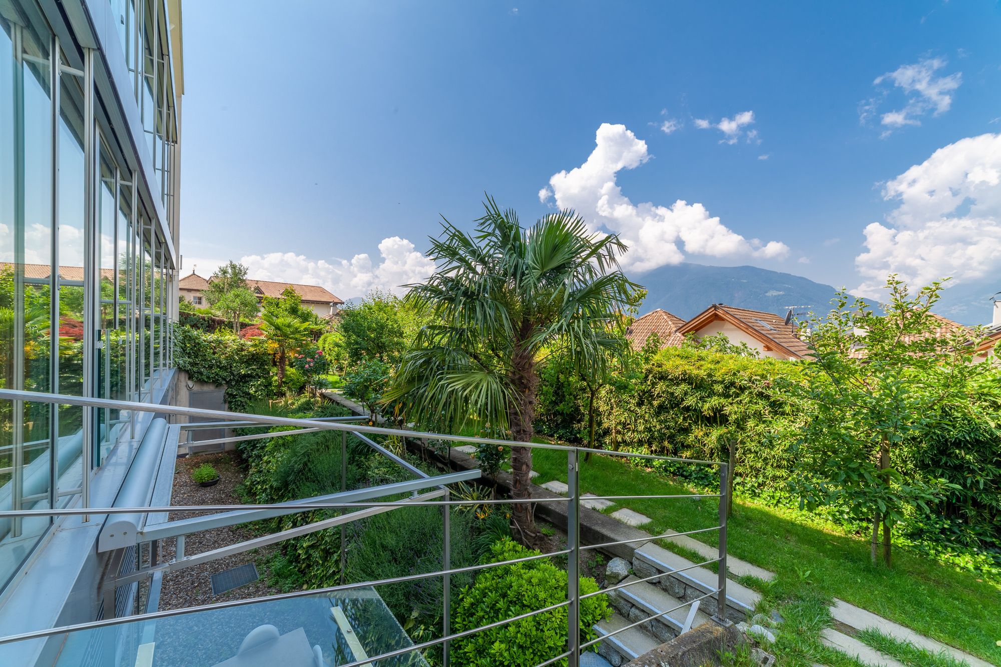 Balkon mit Glasfassade und Geländer, Blick auf grünen Garten mit Palme, Häusern und Bergen unter blauem Himmel. Balkon mit Glasfassade und Geländer, Blick auf grünen Garten mit Palme, Häusern und Bergen unter blauem Himmel.