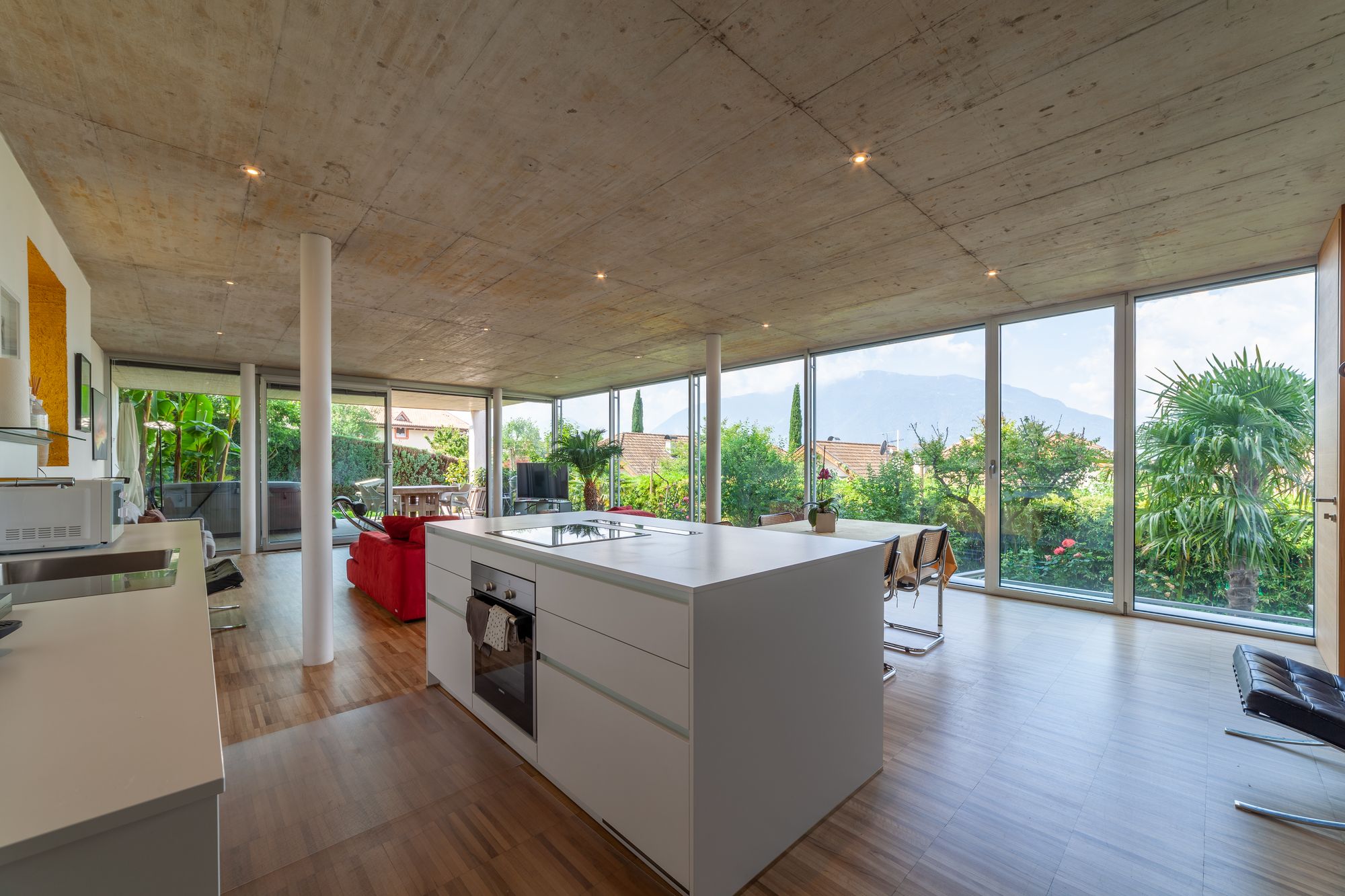 Offener Wohnbereich mit weißer Kücheninsel, roten Sofa und großen Panoramafenstern zum grünen Garten Offener Wohnbereich mit weißer Kücheninsel, roten Sofa und großen Panoramafenstern zum grünen Garten