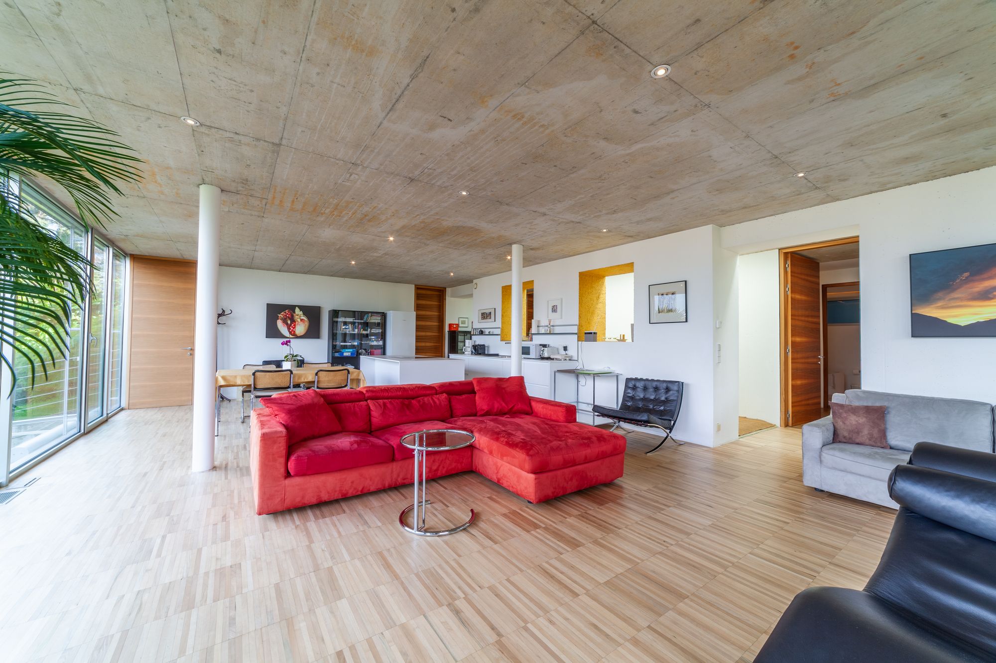 Moderner offener Wohnraum mit rotem Ecksofa, großen Panoramafenstern, Betondecke und Holzfußboden. Moderner offener Wohnraum mit rotem Ecksofa, großen Panoramafenstern, Betondecke und Holzfußboden.