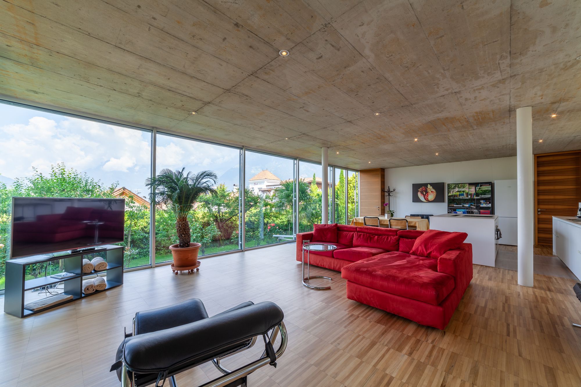 Helles Wohnzimmer mit großen Panoramafenstern, rotem Ecksofa, TV, Topfpflanze und Holzfußboden. Helles Wohnzimmer mit großen Panoramafenstern, rotem Ecksofa, TV, Topfpflanze und Holzfußboden.